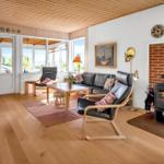 Wohnzimmer mit Holzboden, Sofa, Sessel und Kaminofen. Große Fenster und Tür zum Außenbereich.