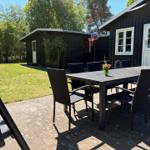 Außenbereich mit Tisch, Stühlen und Grillplatz vor einem schwarzen Holzhaus.