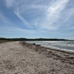 Strand mit Sand und Wellen, Himmel mit weißen Wolken, Ferne mit grünen Hügeln.