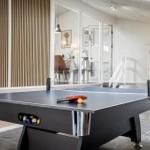 Tischtennisplatte mit Ball und Schläger in einem Raum mit Holzwand und Uhr an der Wand.