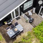 Terrasse mit Grill, Tisch und Stühlen. Schwarze Fassade und Pflanzen umgeben den Außenbereich.