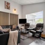 Wohnzimmer mit Sofa, zwei Sesseln, Fernseher und Kaminofen. Große Fenster mit Jalousien.