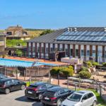 Poolbereich mit Sonnenliegen, Spielplatz und Parkplatz vor einem Gebäude mit Solaranlage.
