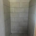 Dusche mit grauen Fliesen und offener Tür zur Baustelle.