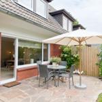 Terrasse mit Tisch, Stühlen und Sonnenschirm. Durchgang ins Haus. Garten mit Hecke und Zaun.