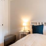 Schlafzimmer mit Bett, Nachttisch und Lampenlicht. Kleiner Sitzhocker und weißer Schrank.