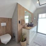 Badezimmer mit Toilette, Waschtisch und Fenster. Holzwand und Spiegel sichtbar.