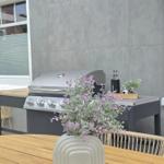 Terrasse mit Grill, Holztisch und Vasenblumen vor grauer Wand.