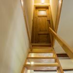 Holztreppe mit Handlauf führt zu einer Holztür. Deckenleuchte beleuchtet den Flur.