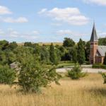 Rotes Kirchengebäude mit Turm, umgeben von Grünflächen und Bäumen unter blauem Himmel.