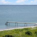 Ein hölzerner Steg führt ins Wasser. Im Hintergrund sind Windkraftanlagen sichtbar.