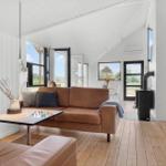 Wohnraum mit braunem Sofa, Holzboden und großen Fenstern. Kamin und TV-Möbel sind sichtbar.