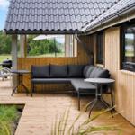 Terrasse mit Sofa, Tischen und Grill unter Dach. Holzwand und Grün im Hintergrund.