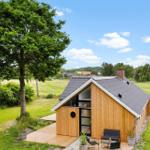Holzhütte mit Terrasse und Garten. Blick auf Felder und Bäume unter blauem Himmel.