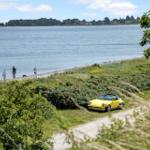 Gelber Sportwagen parkt am Strand. Zwei Personen mit Hund spazieren am Wasser.