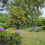 Garten mit Liegestühlen, Blumen und Bäumen. Teil eines Hauses mit Überdachung.