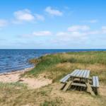 Holzpicknicktisch auf grasigem Hang neben Strand und Meer unter blauem Himmel.