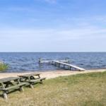 Zwei Holztische stehen am Strand. Hinter ihnen führt eine hölzerne Brücke ins Wasser.