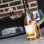 Eine Flasche Wein und ein Glas stehen neben einem Grill auf einer Terrasse.
