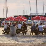 Besucher sitzen unter roten Sonnenschirmen an Tischen am Hafen.