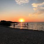 Sonnenuntergang am Strand mit Menschen im Wasser und Sandstrand im Vordergrund.