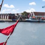 Rotes Segel an einem Mast vor einem Hafen mit Booten und Häusern.