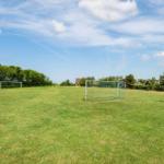 Grünes Spielfeld mit Fußballtor und Basketballkorb unter blauem Himmel.