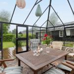 Glasveranda mit Holztisch, Stühlen und Blumen. Draußen ist ein Garten mit Rasen und Bäumen.