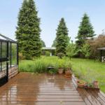 Holzterrasse mit Pflanzen, Gartenhaus und Pavillon im Hintergrund.