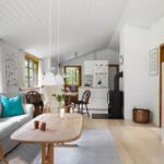 Wohnzimmer mit Sofa, Tisch und Küchenbereich. Holzdielen, weiße Wände und Fenster mit Holzrahmen.