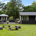 Schwarze Holzhütte mit Garten, Tisch, Bänken und Sonnenschirm. Umgeben von Bäumen.