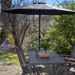 Terrasse mit Tisch, Stühlen und Sonnenschirm. Hintergrund mit Bäumen und Sträuchern.