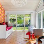 Kinderzimmer mit buntem Wandbehang, Schlafcouch, Spielzeug und großem Fensterblick auf Garten.