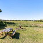 Holztisch und Grillplatz auf grünem Rasen mit Blick auf Feld und Bäume.