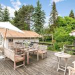 Holzterrasse mit Tisch, Bänken und Sonnenschirm. Hintergrund: Wald und Garten.