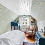 Schlafzimmer mit gemustertem Tapeten, Bett, Sessel und Schrank unter Dachschrägen.