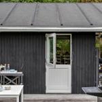 Eine schwarze Hütte mit weißer Tür, Terrasse mit Tisch, Blumen und Grill.