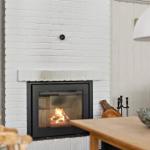 Kamin mit brennendem Feuer, weiß verputzte Mauer, Holztisch und Stuhl im Vordergrund.