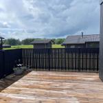 Holzterrasse mit schwarzer Umzäunung und Gartenmöbeln. Im Hintergrund sind Häuser und Wiesen zu sehen.
