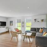 Wohnzimmer mit Sofa, zwei Sesseln, Couchtisch und Fernseher. Holzboden und große Fenster.