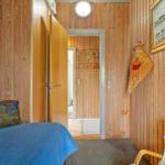 Schlafzimmer mit Holzwand, Bett, Stuhl und Wandbild. Tür führt in einen Flur.