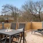 Terrasse mit Tisch, Stühlen und Liegen. Holzgeländer und Bäume im Hintergrund.