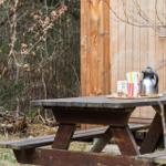 Holztisch im Freien mit Kanne, Tassen und Flasche vor Holzwand und Wald.