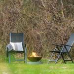 Zwei Gartenstühle und ein Feuerkorb stehen auf einer Wiese vor Sträuchern.