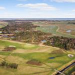Luftbild eines Golfplatzes mit umliegenden Wiesen und Bäumen. Im Hintergrund ist ein Gewässer zu sehen.