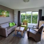 Wohnzimmer mit Sofa, Sessel und TV. Große Fenster mit Blick auf grüne Hecke.