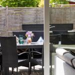 Terrasse mit Tisch, Stühlen und Grill. Hintergrund: Holzzaun und grüne Bäume.