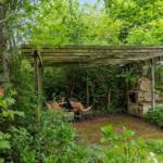 Gartenbereich mit Holzüberdachung, Sitzgelegenheiten und Steinofen zwischen Bäumen.
