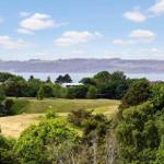 Weitläufiger Golfplatz mit Blick auf Wasser und Hügel unter blauem Himmel.