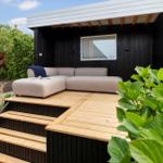 Terrasse mit grauem Sofa und Holzstufen vor schwarzem Holzhaus mit Fenster.